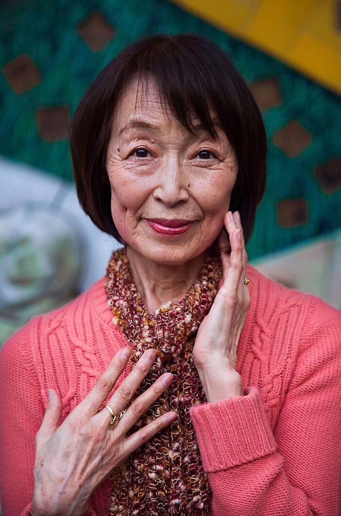 Elderly woman with short dark hair wearing a pink sweater and scarf, smiling gently in a colorful outdoor setting.