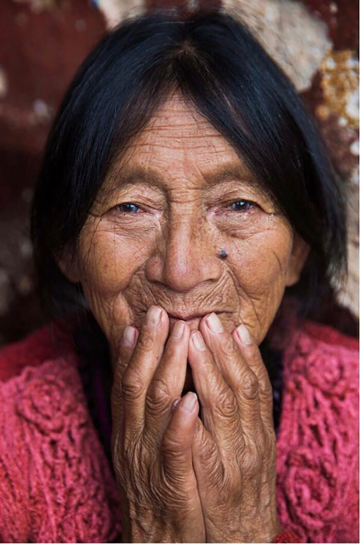 Elderly woman with dark hair and textured skin smiling gently, hands covering mouth, showcasing timeless beauty and wisdom.
