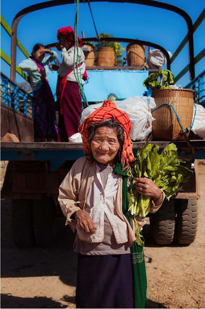 Elderly woman wearing a red headscarf holding fresh greens, standing in front of a truck with baskets behind her.