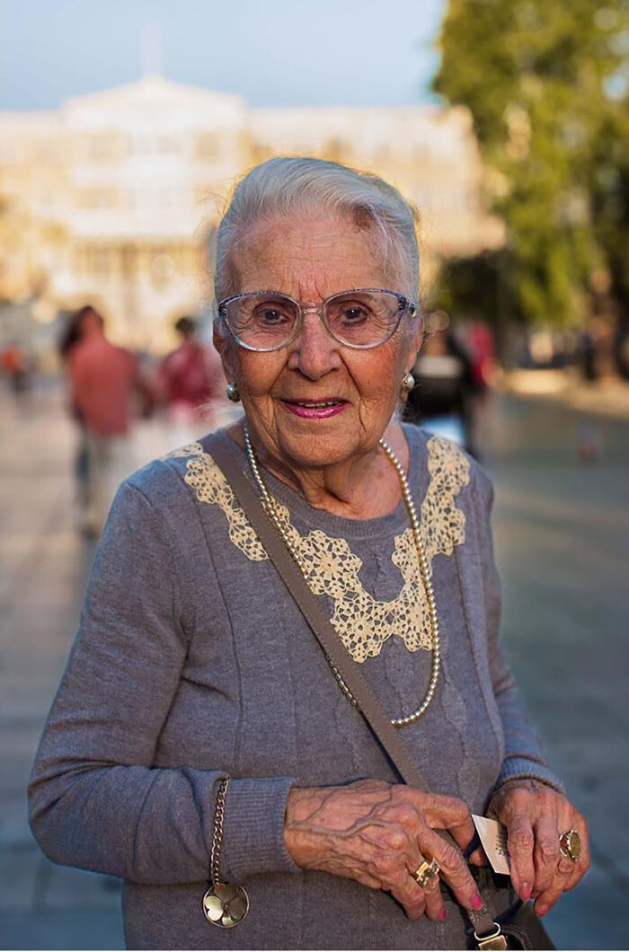 Elderly woman wearing glasses and pearl necklace smiling outdoors, showcasing beauty and grace of elderly women worldwide.