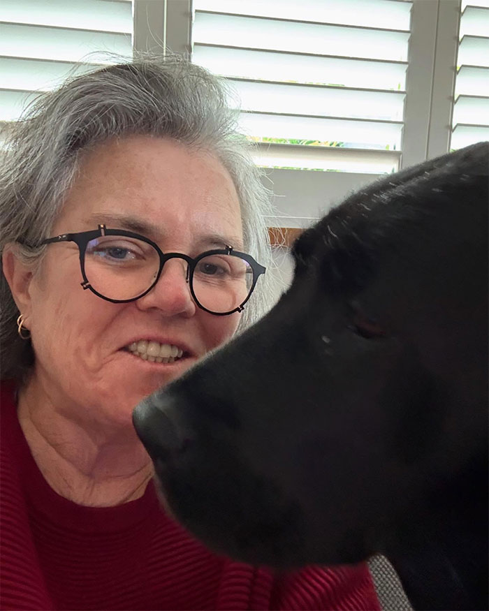 Woman with glasses smiling next to a black dog indoors, representing stars who said goodbye to the US and found new home abroad.