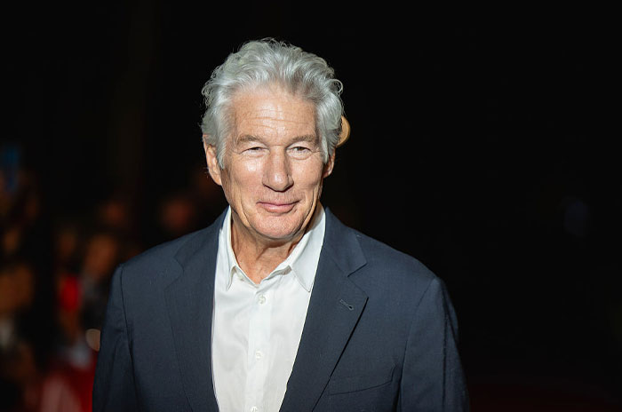 Richard Gere at an event, dressed in a dark blazer and white shirt, representing stars who found new homes abroad.