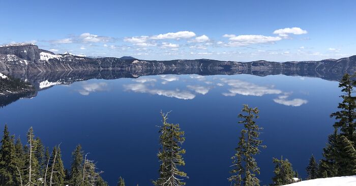 Calm blue lake reflecting clouds and mountain cliffs, surrounded by evergreen trees in a breathtaking travel photo.
