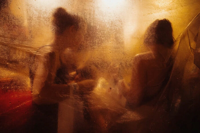 Blurry candid street photo showing two people sitting behind a textured, translucent surface in warm lighting.