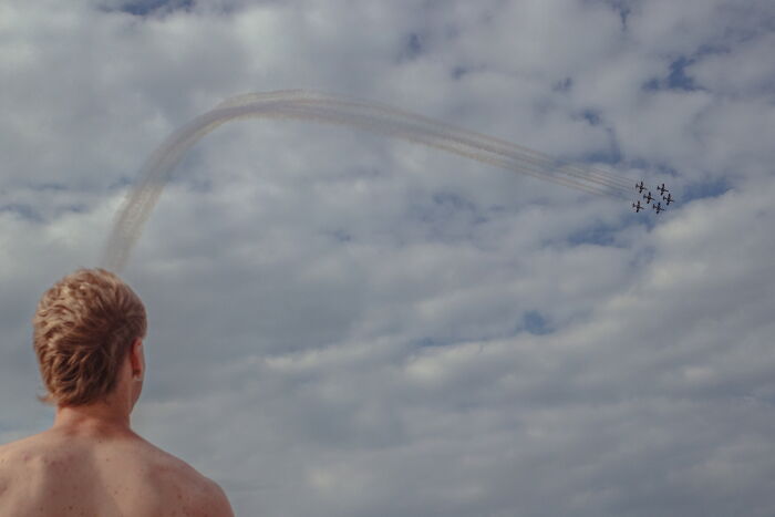 Shirtless man watching perfectly-timed street photo of airplanes creating a curved smoke trail across a cloudy sky.