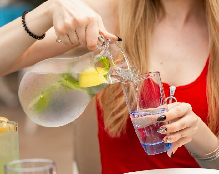 Person with long hair in a red top pouring water from a pitcher with lemon and mint into a glass, illustrating poor people habits.