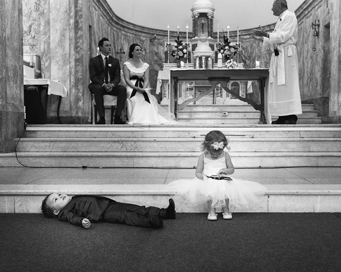 Black and white wedding photo capturing candid moments with bride, groom, children, and priest during ceremony in church.