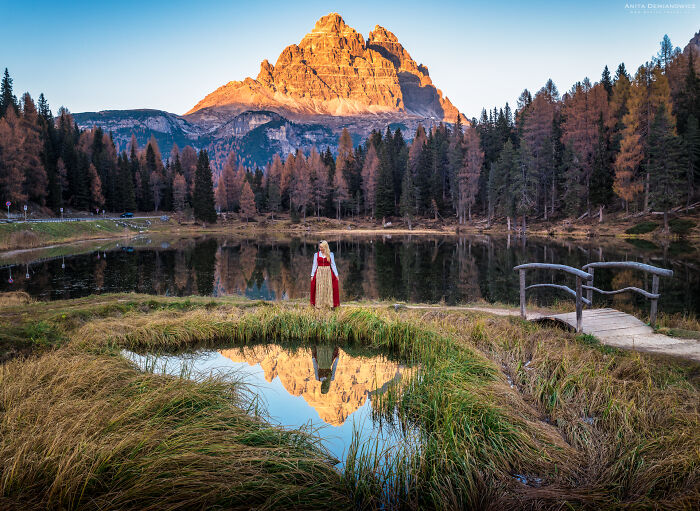 Woman in traditional dress by a mountain lake reflecting autumn trees at sunset, breathtaking travel photo captured outdoors.