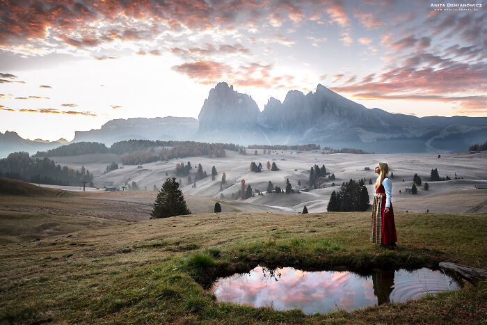 Woman in traditional dress standing near a pond with mountains and a colorful sky in breathtaking travel photos scenery.