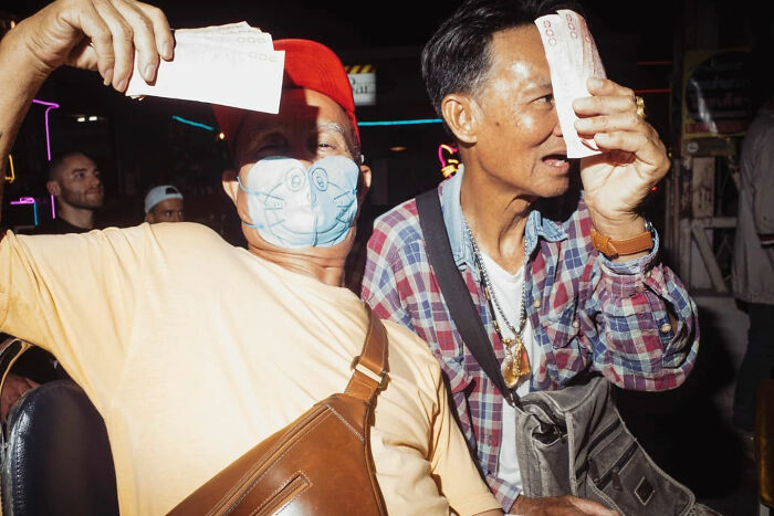 Two men at night market, one wearing cartoon face mask, holding money in candid street photos by Alex McClintock.