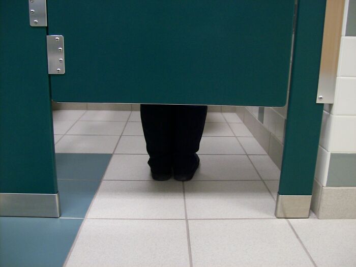 Person standing behind a bathroom stall door with only feet visible, illustrating human behavior and personal mysteries.