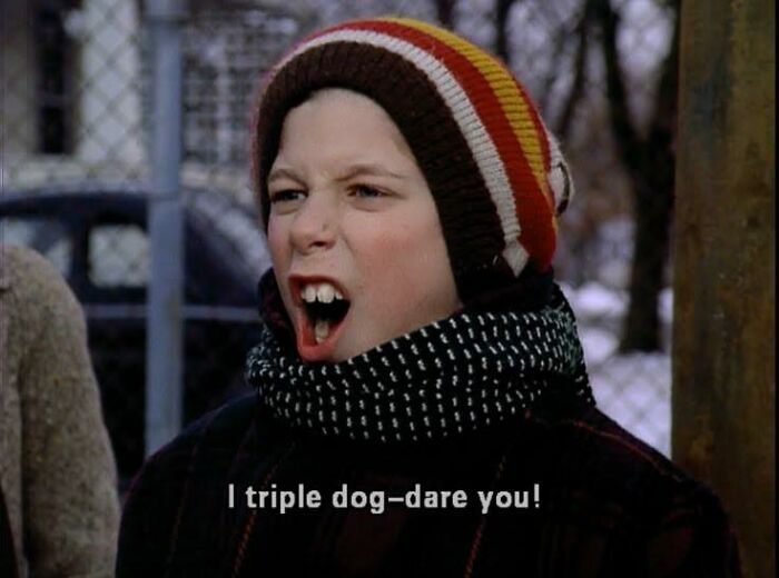 Young boy wearing a striped beanie and scarf, shouting the phrase triple dog-dare you outdoors in winter setting.