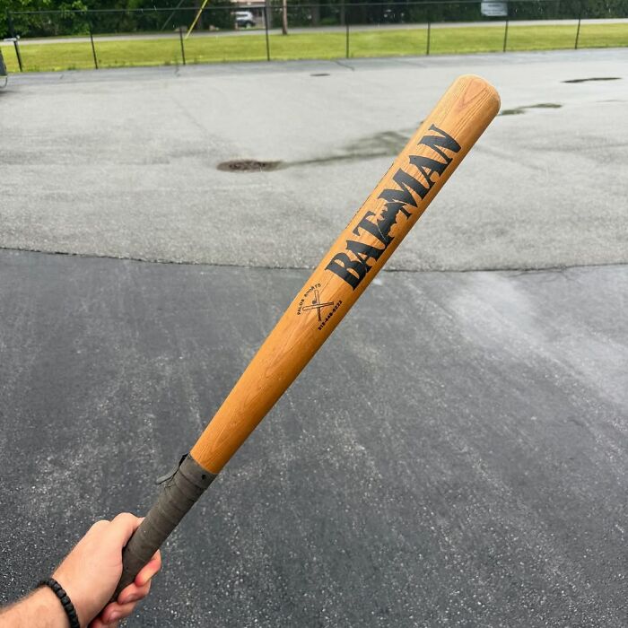 Hand holding a secondhand wooden baseball bat with Batman logo outdoors on an asphalt surface.