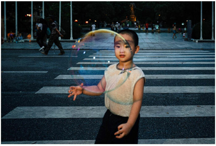Young boy playing with large bubble on a busy street, capturing raw and captivating moments from the streets of Asia.
