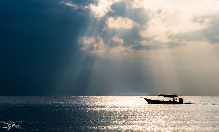 Silhouette of a small boat on shimmering water under dramatic clouds with sun rays, a breathtaking travel photo.