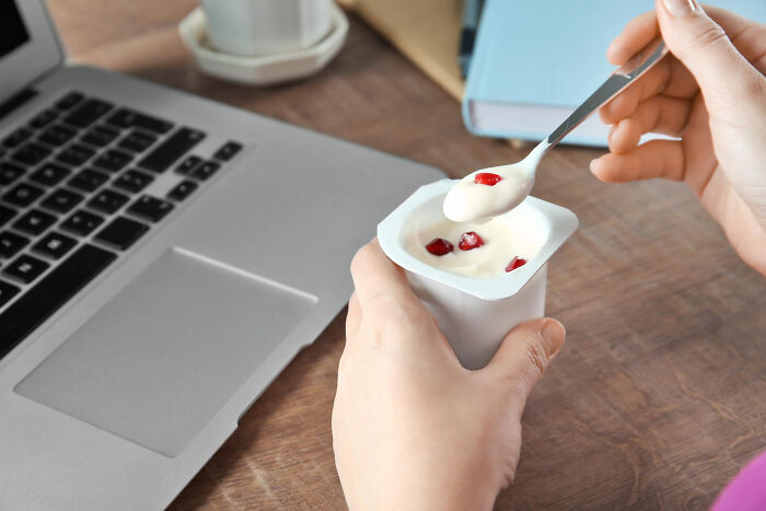 Person eating yogurt with a spoon near a laptop, illustrating poor people habits in daily routines and lifestyle choices.