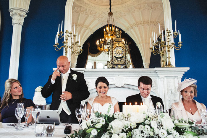 Bride and groom reacting during wedding speech at an elegant venue with floral arrangements and candles in front.