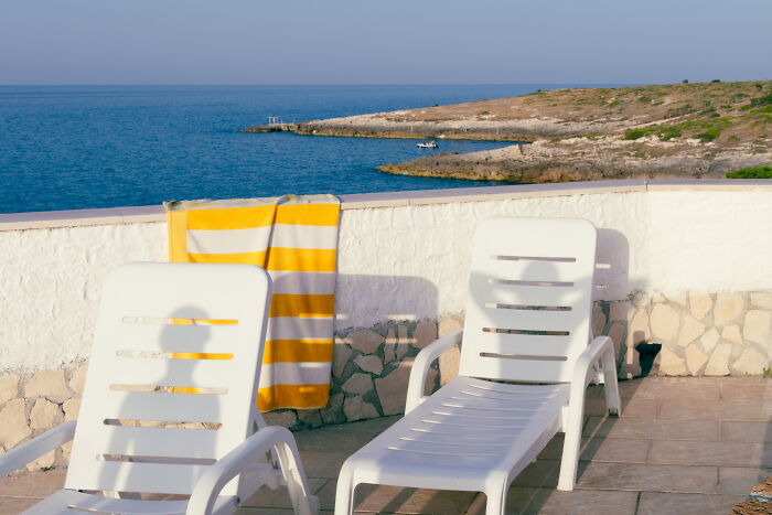 Two white lounge chairs with a yellow striped towel on a stone patio overlooking the sea in a street photo.