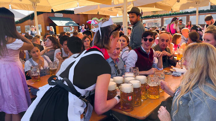 A crowded outdoor beer garden with people enjoying drinks and a waitress serving large mugs European netizens common facts