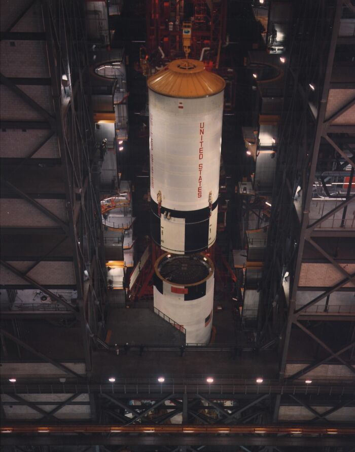 Apollo mission rocket stages being assembled inside a large spacecraft hangar with workers and structural beams visible.