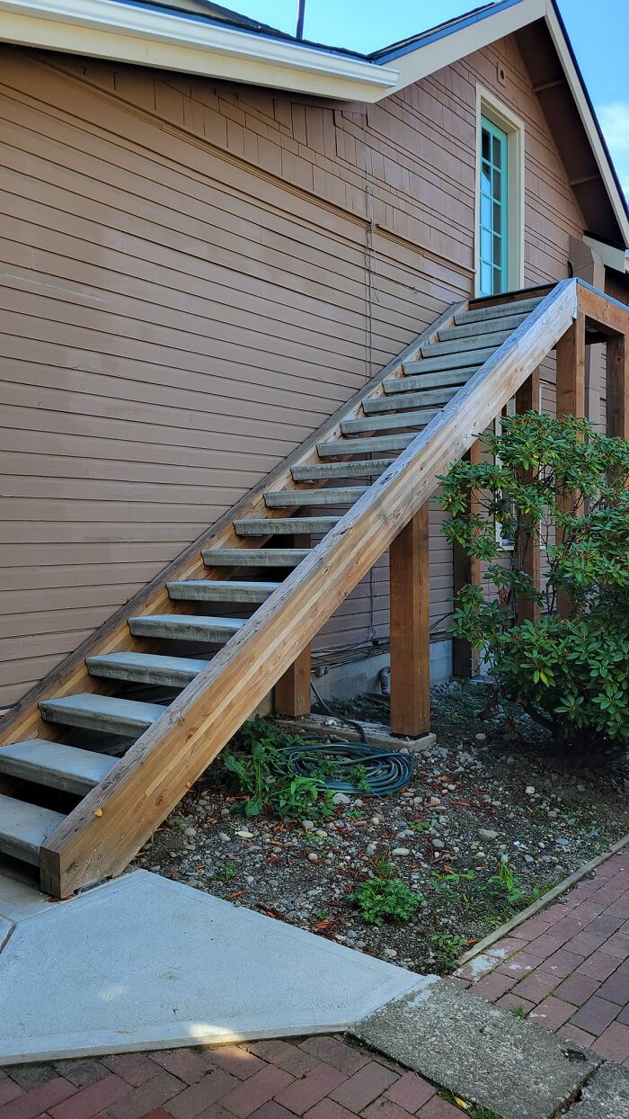 Steep wooden outdoor death stairs with narrow steps attached to house, surrounded by plants and a brick walkway.