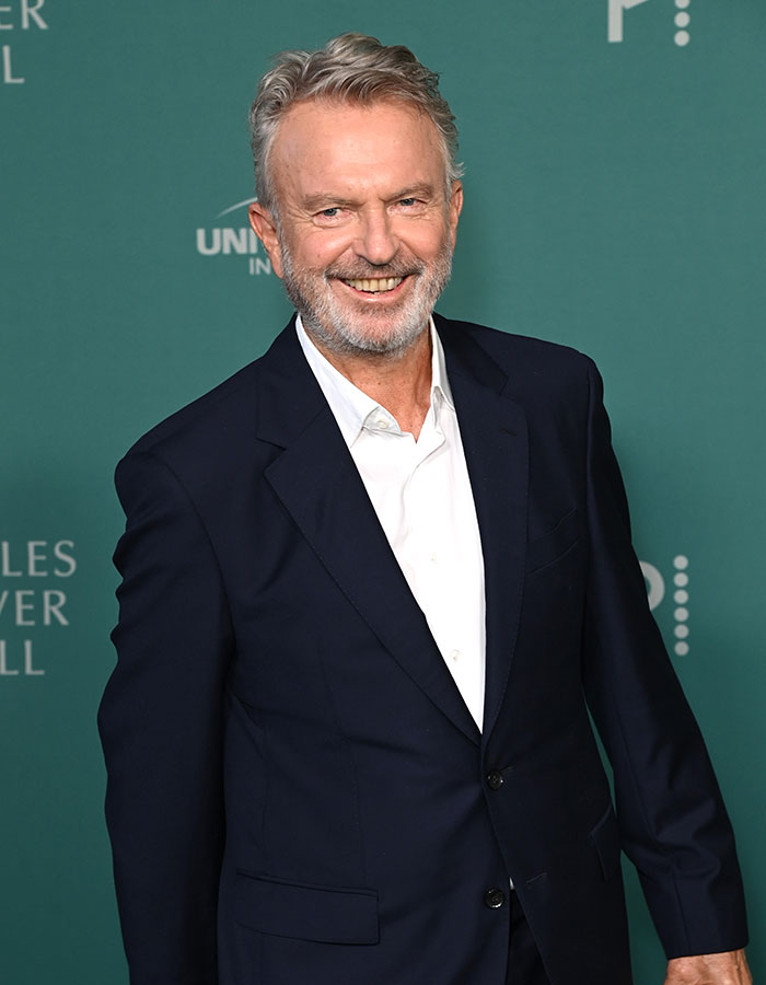 Older male celebrity with gray hair smiling, dressed in a dark suit and white shirt, showing youthful appearance among celebrities.