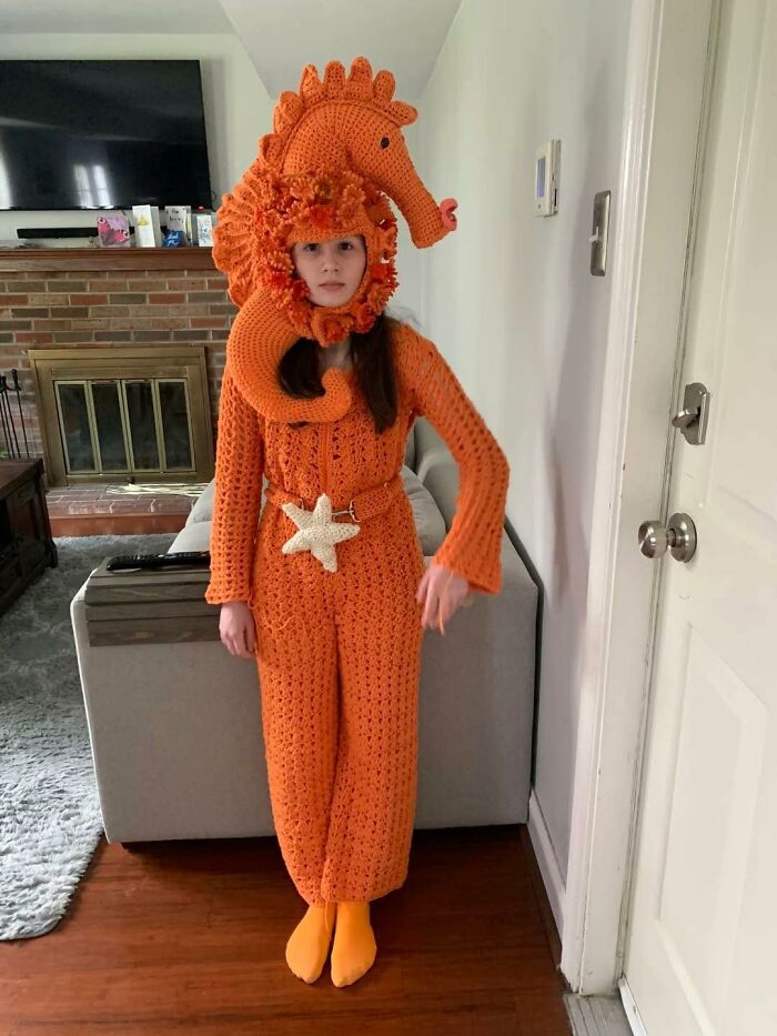 Girl wearing an orange crocheted seahorse costume standing indoors, an example of horrible crafts that netizens shamed online.