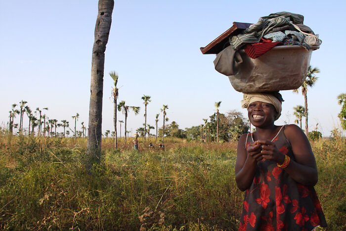Smiling woman carrying a basket on her head in a rural field with palm trees, showcasing breathtaking travel photos.
