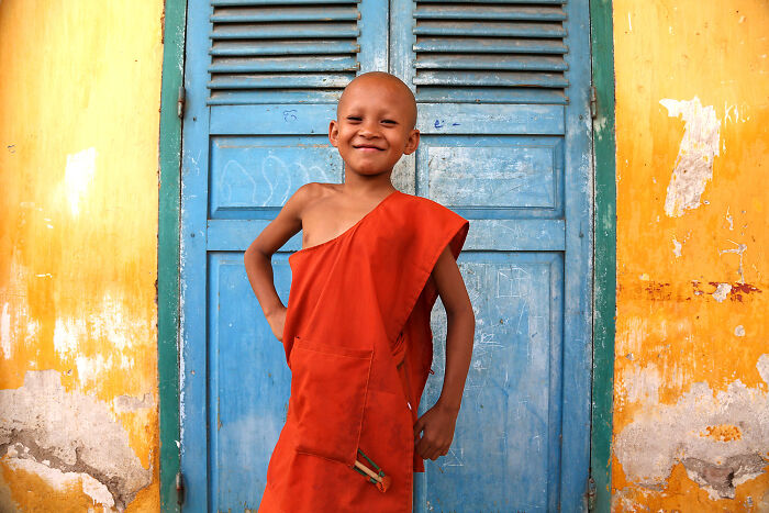 Young monk smiling confidently in vibrant orange robes against colorful blue and yellow walls, a breathtaking travel photo.