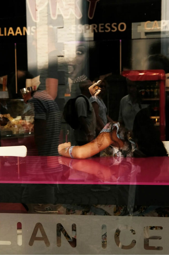 Reflection of people walking past a cafe window with a woman resting on a vibrant pink table in this beautiful street shot.