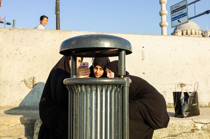 Three women in black hijabs taking a selfie behind a trash can, showcasing street photographer catching the world off guard.