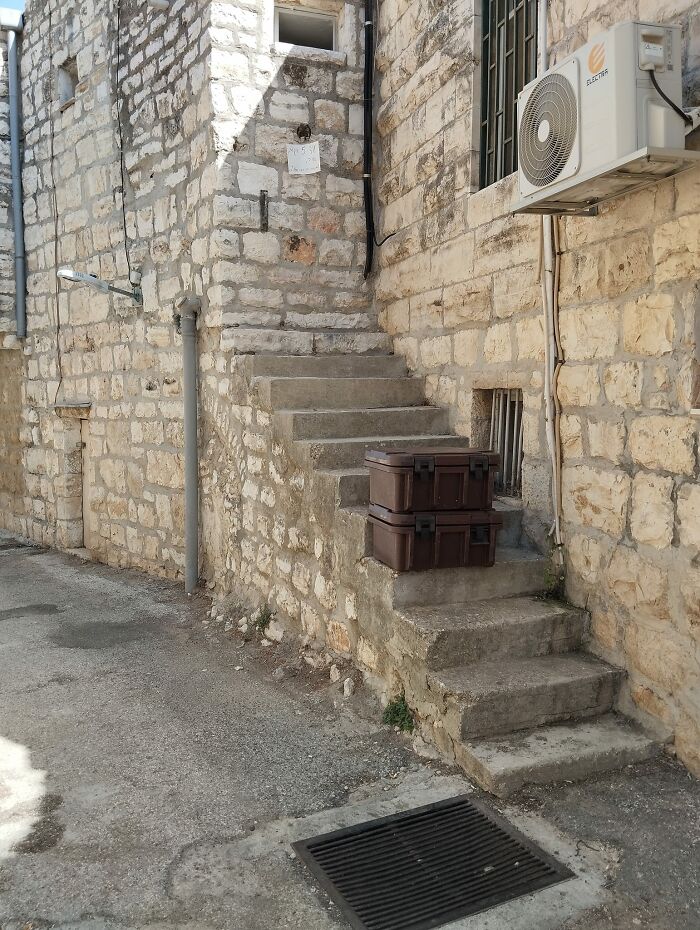 Narrow stone death stairs blocked by two stacked boxes, attached to an old stone building with an air conditioner unit.