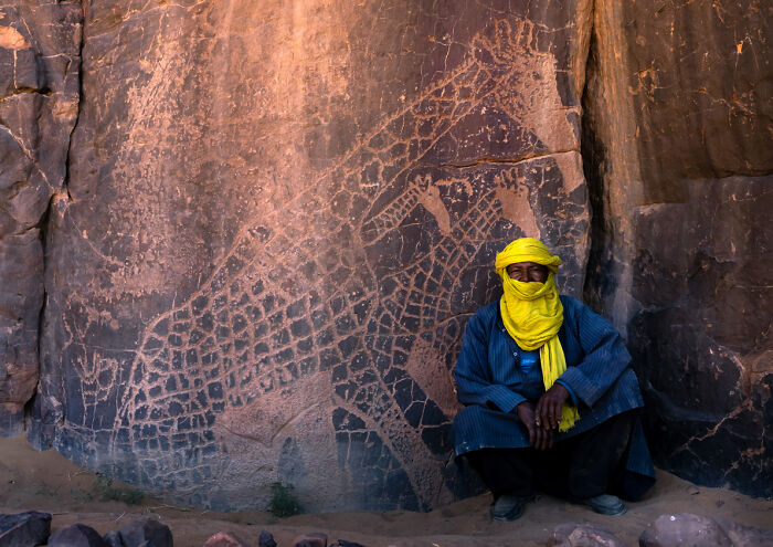 Man in traditional wear sitting by ancient giraffe rock carvings, a breathtaking travel photo from Bored Panda community.