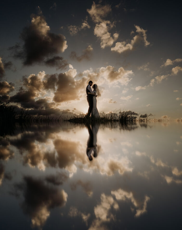 Silhouette of a bride and groom embracing at sunset with dramatic clouds and a reflective water surface in unforgettable wedding photos.