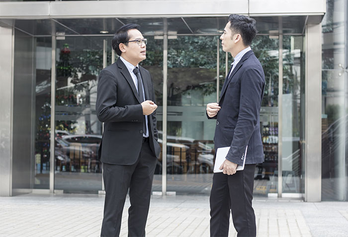 Two businessmen in suits discussing outside a modern building, highlighting an insurance company denying employee claim. Two businessmen in suits discussing outside a modern building, highlighting an insurance company denying employee claim.