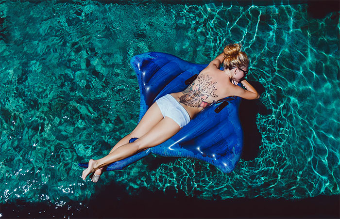 Woman sunbathing topless on a blue inflatable float in a pool, showing a large back tattoo under the sunlight.