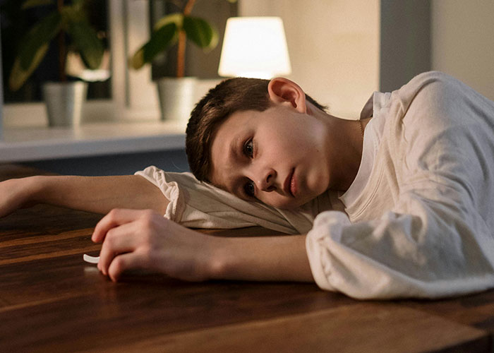 Boy lying on wooden table with tired expression, capturing a moment of taking one for the team in a quiet indoor setting.