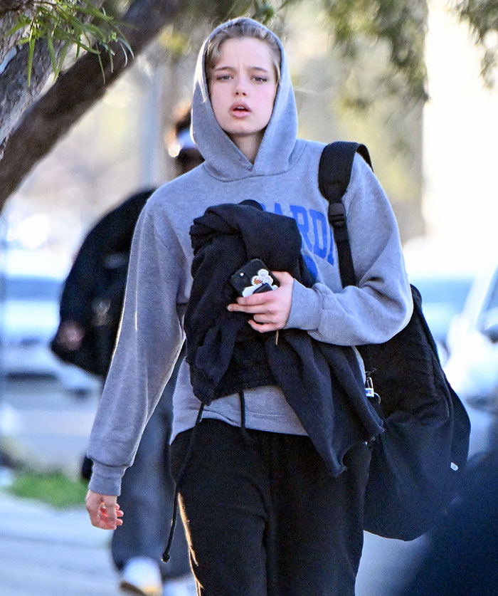 Young person in a grey hoodie walking outdoors, holding clothes and a phone, linked to Brad Pitt cutting off contact with sons news.