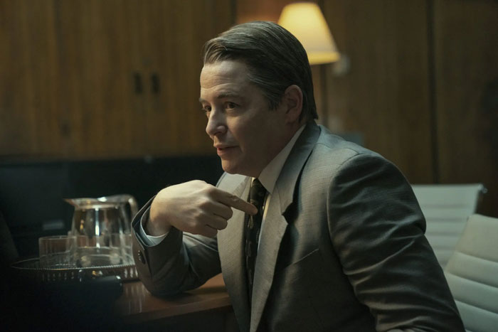 Man in gray suit and tie talking about famous people who did evil things in the past in a dimly lit office setting.