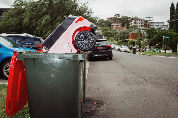 Candid street photo of a toy car stuck in a garbage bin on a suburban street by Alex McClintock.