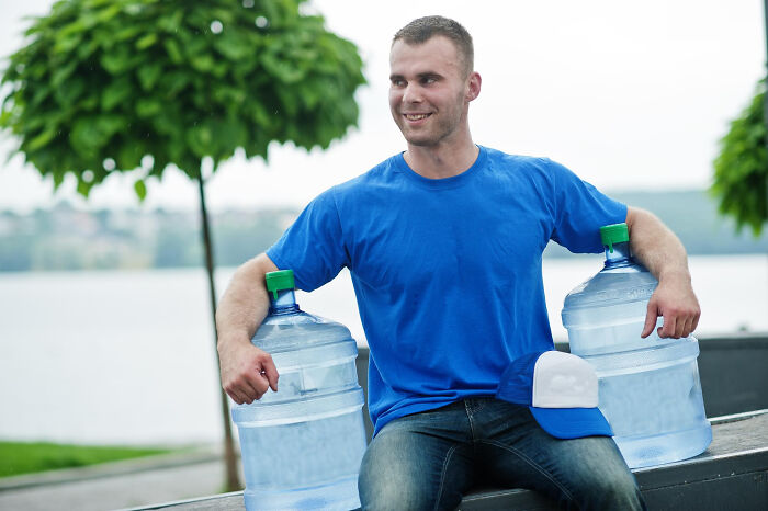 Young man in blue shirt holding two large water containers outdoors near trees and water, symbolizing completely legal total psychopath concept.