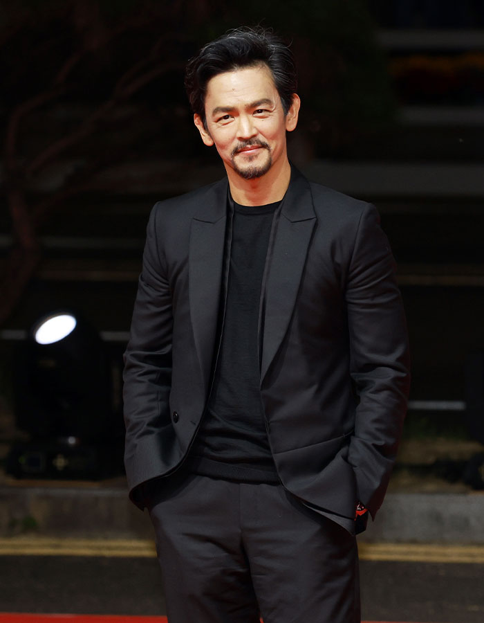 Actor in a black suit smiling confidently, showcasing a youthful appearance among celebrities who look younger than their age.