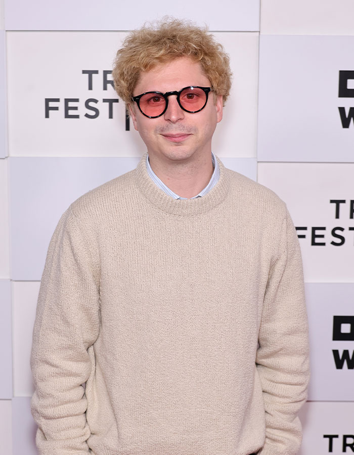 Young-looking celebrity wearing round tinted glasses and a beige sweater at a festival event, showcasing youthful appearance.