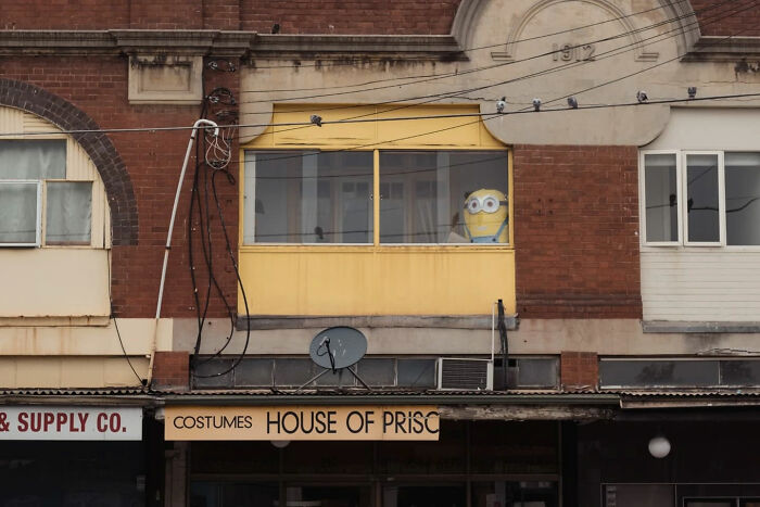 Candid street photo of a yellow brick building with a Minion figure visible inside a second-floor window.