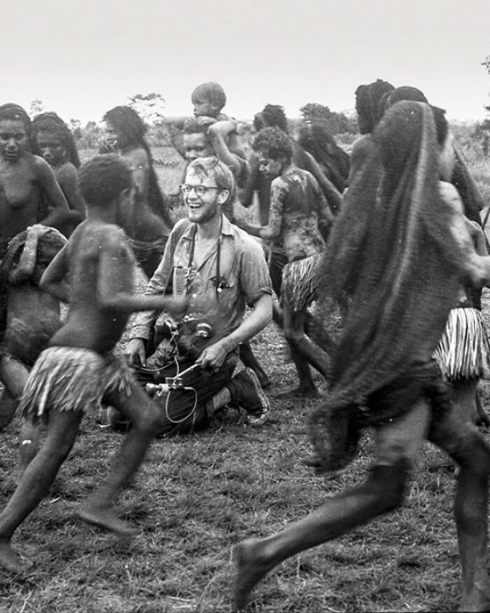 Black and white historic photo showing a group of children playing around a man with a camera in a rural setting.