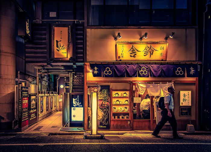 Nighttime street scene of a warmly lit Japanese restaurant with a person walking by in a breathtaking travel photo.