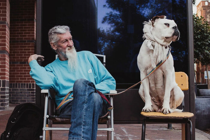 Elderly man with a long beard sitting next to a large white dog on a chair in candid street photos.