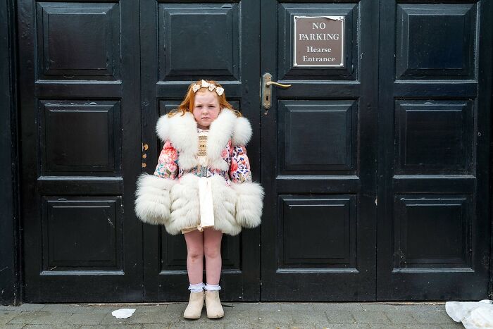 Young girl in a fur-trimmed coat standing solemnly in front of a black door, reflecting life on the margins of modern Ireland.