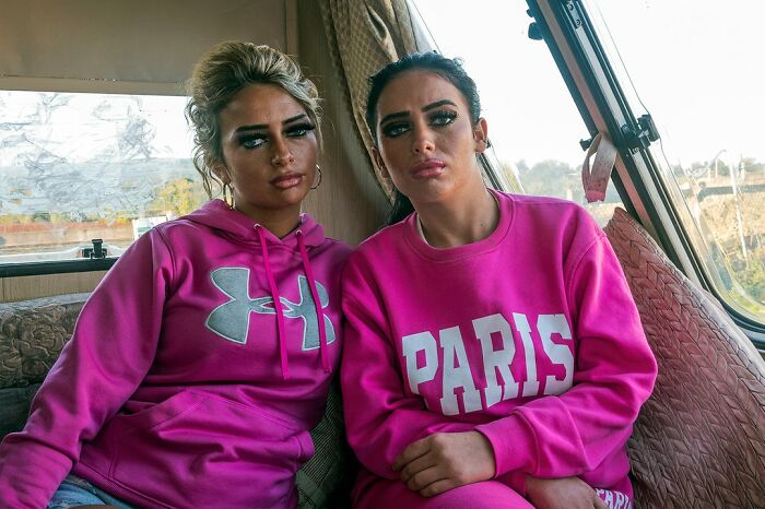 Two young women wearing bright pink sweatshirts, sitting closely inside a vehicle, capturing life on the margins of Ireland.