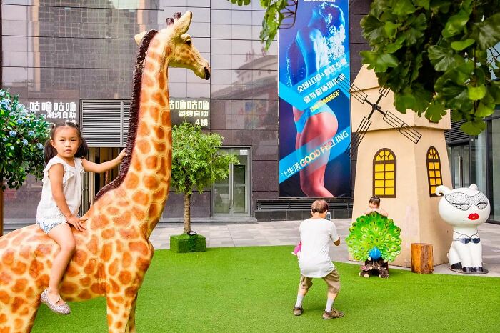 Child riding a giraffe statue in a vibrant urban park captured by award-winning candid street photographer.